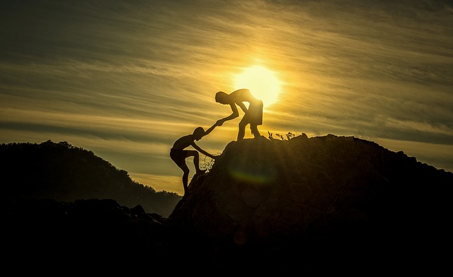 Symbolizing teamwork, one boy helps another navigate a rocky ledge -- all in silhouette against a setting sun.